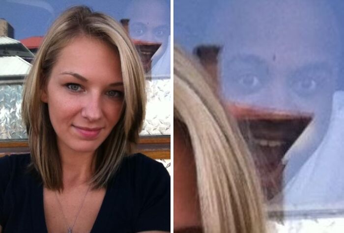 Woman taking a selfie with a photobomb visible as a scary face reflected in the glass behind her during an outdoor moment.