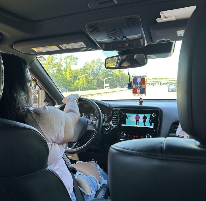 Woman driving a car with torn jeans, watching a video on the dashboard screen, illustrating mildly infuriating things testing patience.