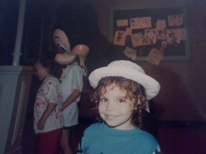 Child smiling at camera in a white hat while a funny photobomb with a character costume happens in the background.