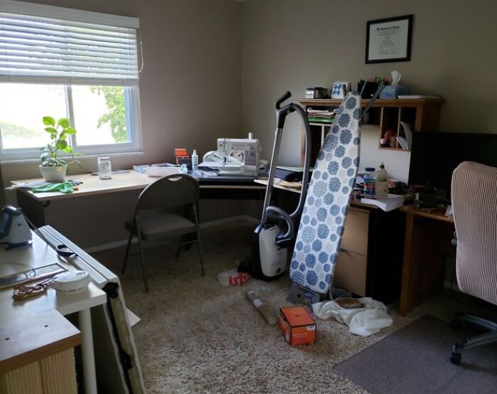 Home office with an ironing board, vacuum, cluttered desk, and a cat seamlessly merged into the background on the floor.