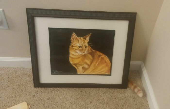 Framed painting of an orange cat blending into carpet with a real cat’s striped tail peeking from behind the frame.
