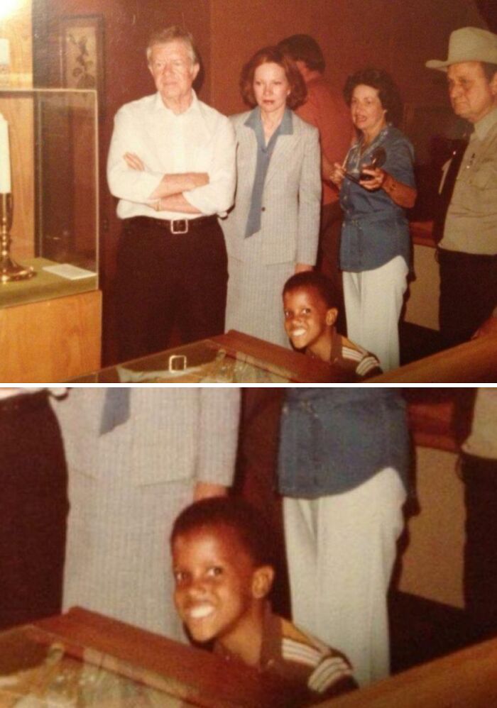 Vintage photo showing a child photobombing adults in a museum, smiling widely behind a display case.