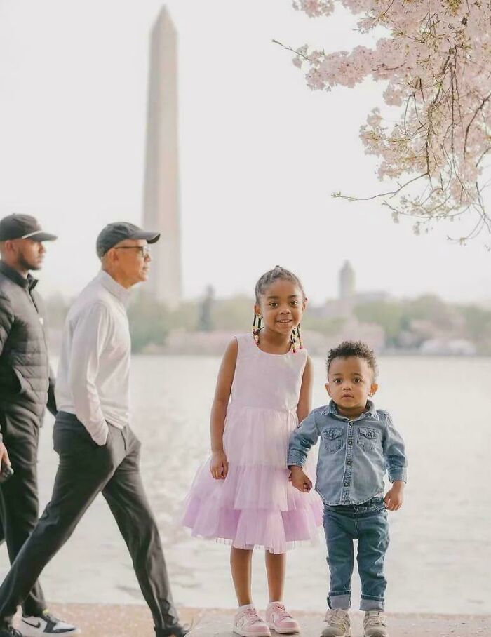 Two children posing by the water with Washington Monument in background while two men photobomb the picture.
