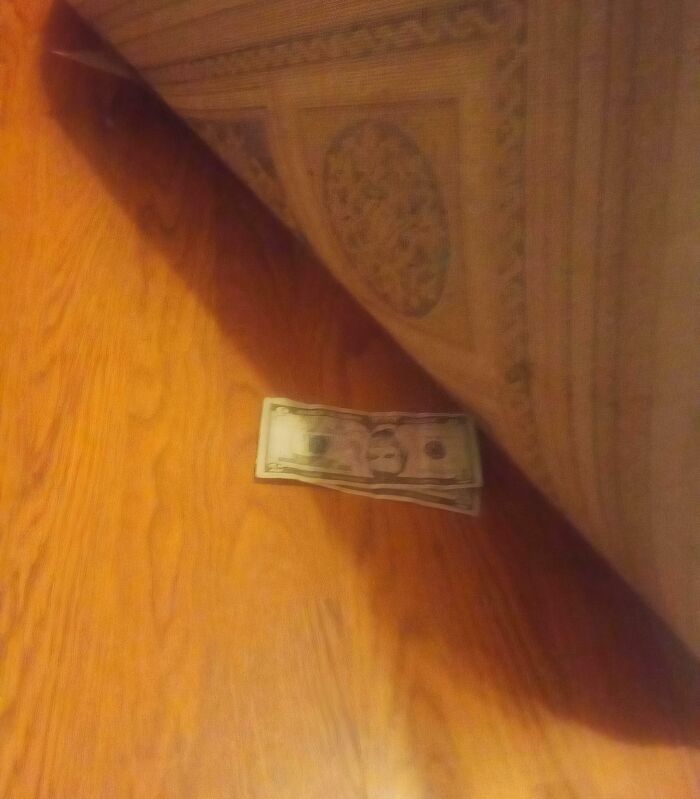 Dollar bills partially stuck under the edge of a rug on a wooden floor, a mildly infuriating thing testing patience.