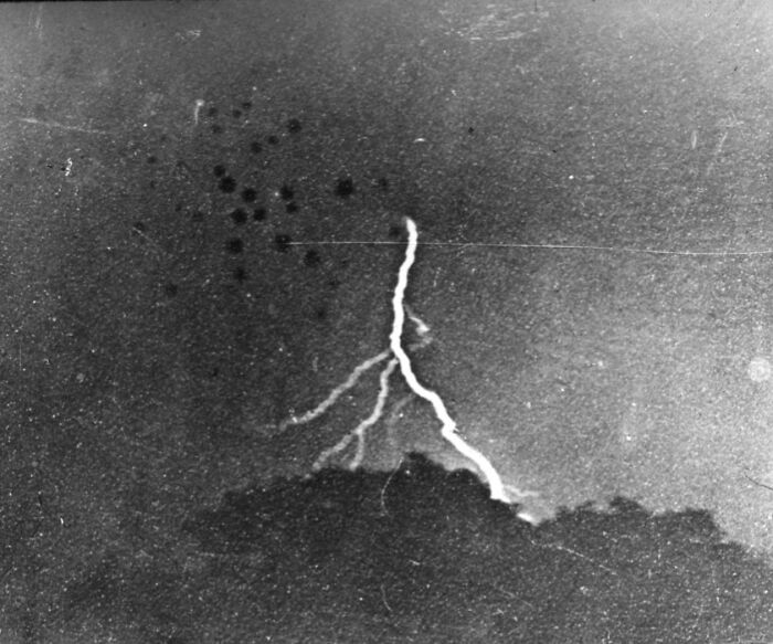 Black and white photo showing early photography capturing lightning striking over dark tree silhouettes at night.