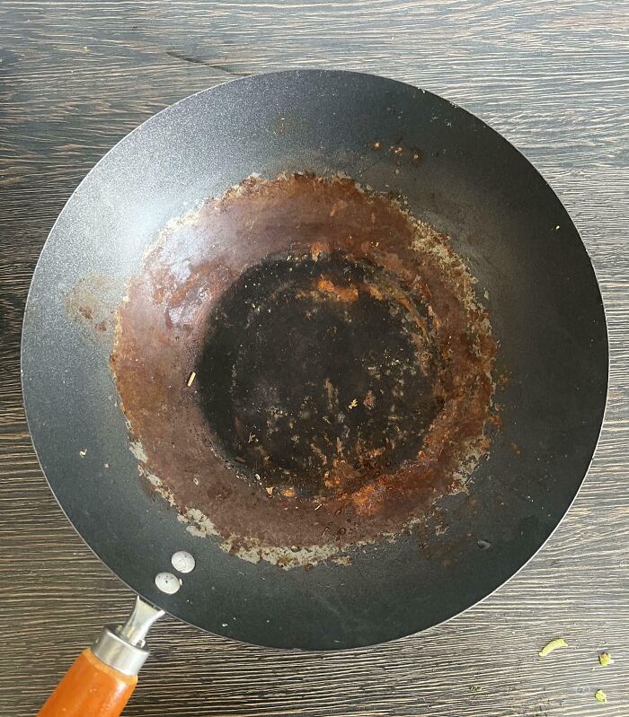 Rusty and burnt frying pan with a wooden handle on a wooden surface, showing mildly infuriating wear and stains.