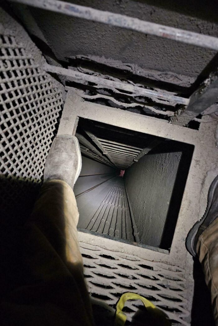 View looking down a narrow, dark industrial shaft with a worker's dusty boot on a metal grate ladder edge.