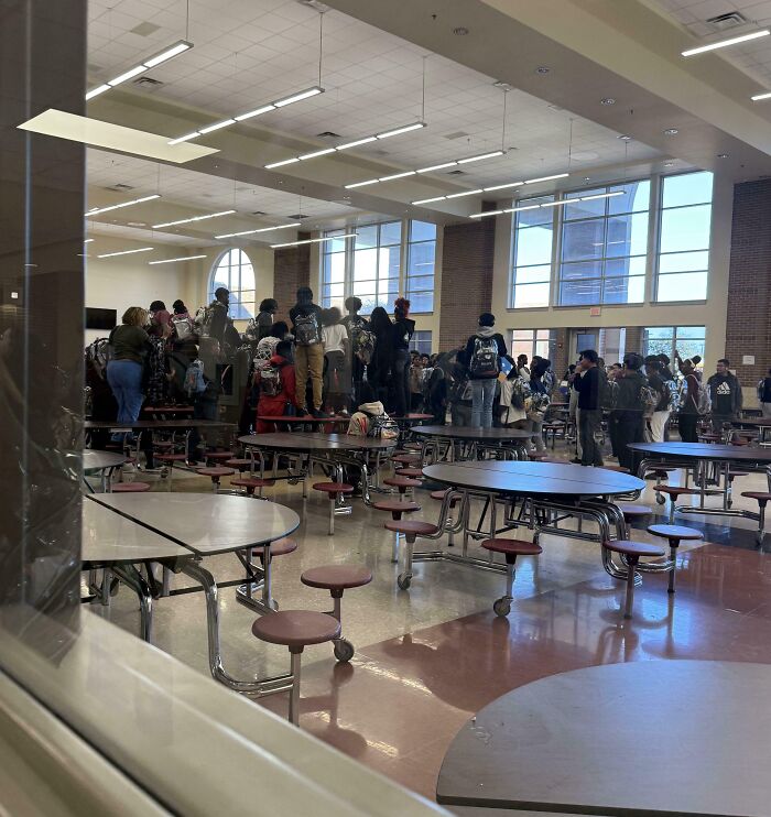 Crowded school cafeteria with students standing on tables, a mildly infuriating scene testing people’s patience.