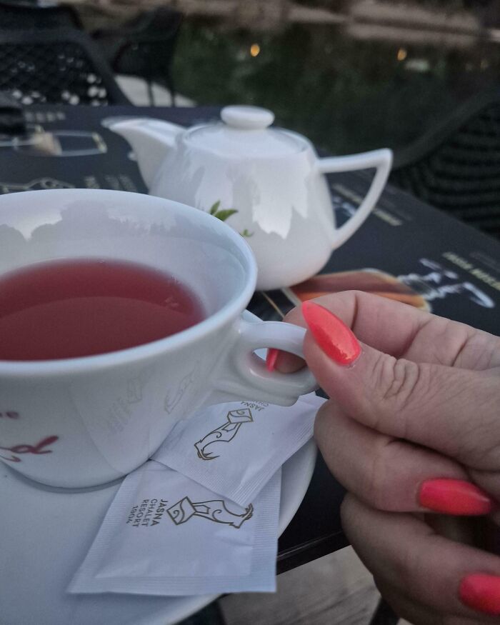 Hand with bright pink nail polish holding a teacup with tea next to sugar packets and a teapot, mildly infuriating scene.