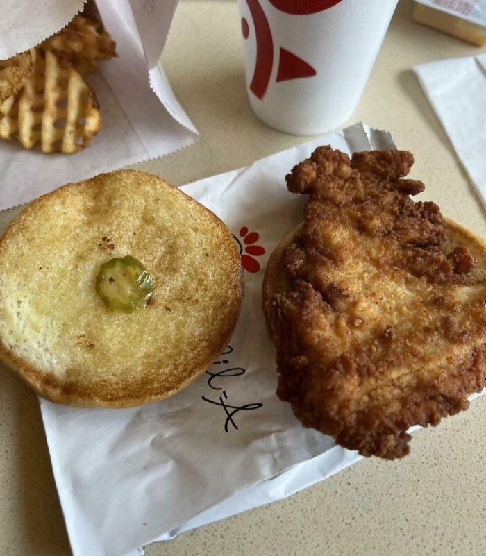 Half-eaten fast food chicken sandwich with only one tiny pickle slice on toasted bun, waffle fries and drink nearby.