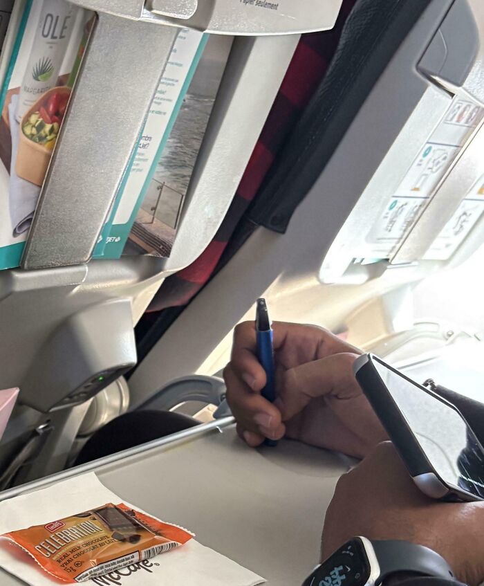 Passenger struggling to write on an airplane tray table, showing a mildly infuriating moment testing patience in travel.