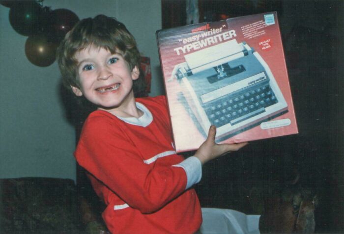 Young boy in a red shirt smiling widely while holding a vintage easy-writer typewriter box from the 1980s era.