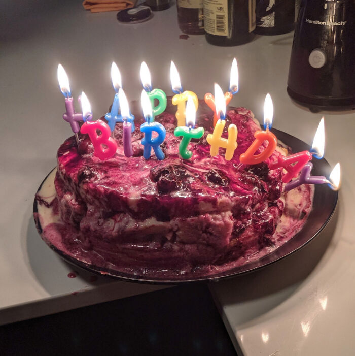 Melting birthday cake with colorful candles and a messy, unappetizing appearance in a kitchen setting.