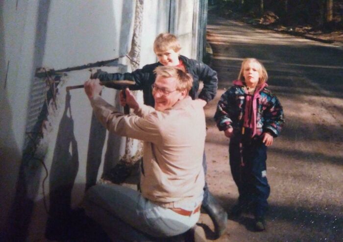 Man in 1980s clothing using a tool on a wall outdoors while two children look on on a sunny day