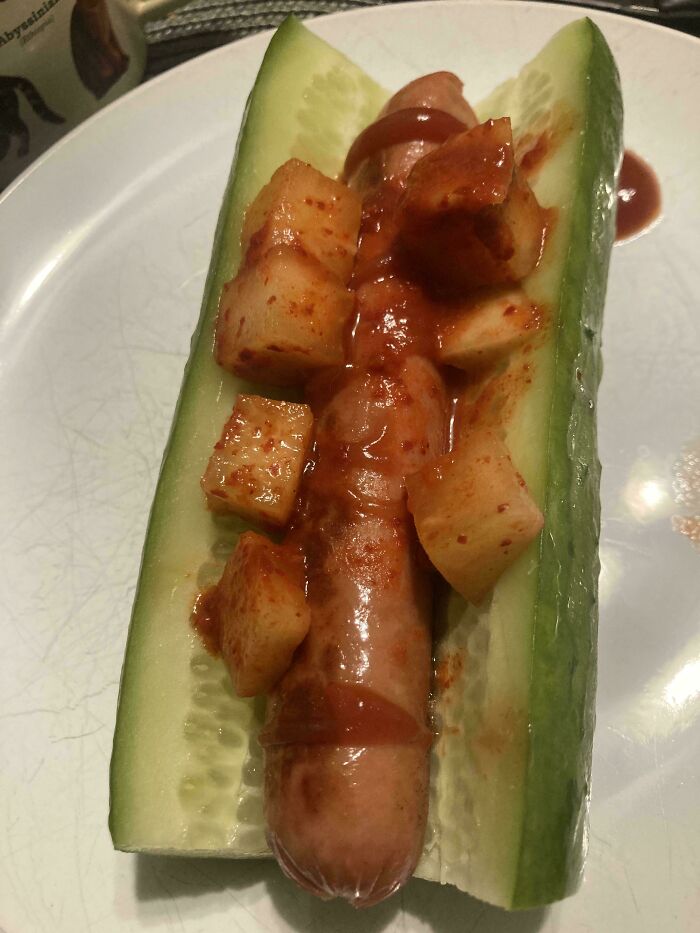 Cucumber sliced in half serving as a bun for a sausage topped with spicy cubed food in a cursed and disgusting food picture.