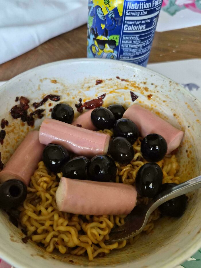 Bowl of instant noodles topped with black olives and sliced hot dog pieces, an example of cursed and disgusting food combinations.