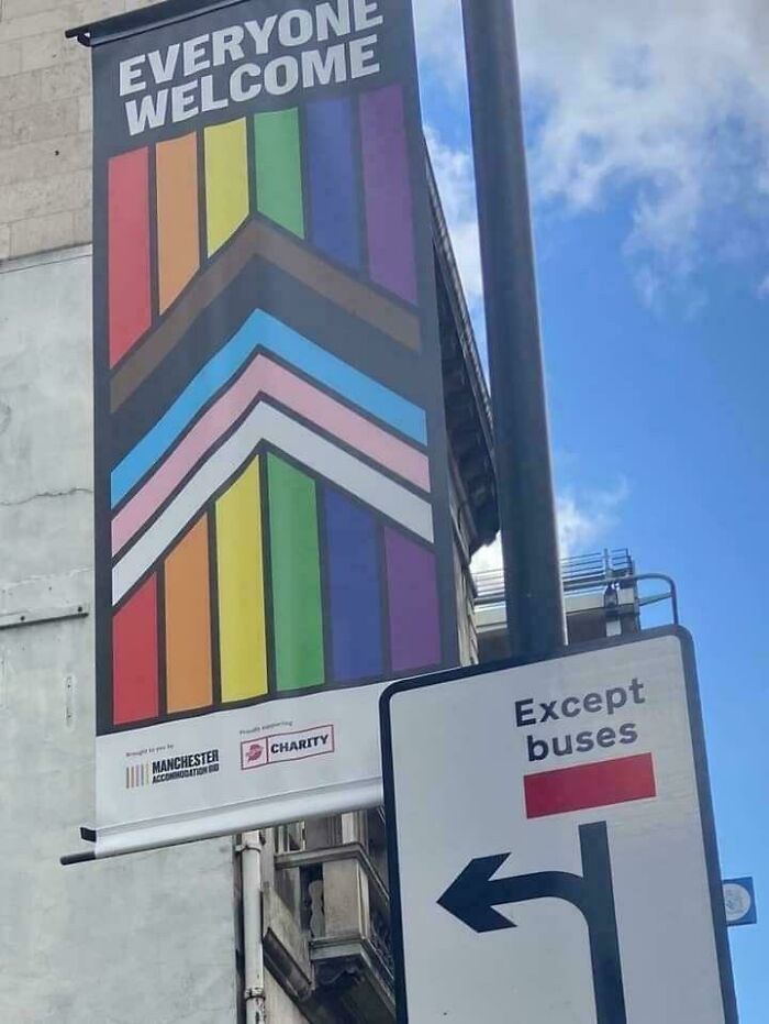 Colorful pride flag banner saying everyone welcome next to a traffic sign allowing left turns except buses in an urban area.