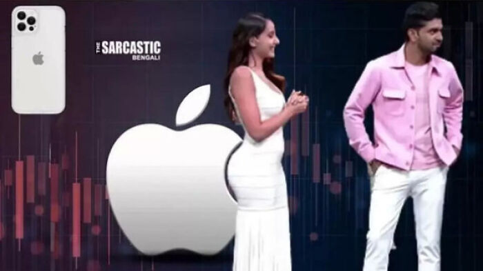 Woman in white dress and man in pink jacket posing in front of a large Apple logo in a scene of accidental comedy.