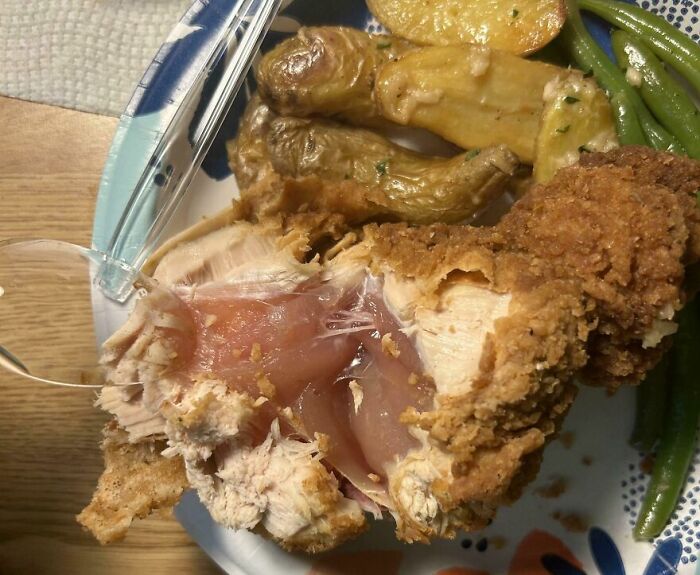 Partially cooked fried chicken with raw pink meat exposed, served with roasted potatoes and green beans on a plate.