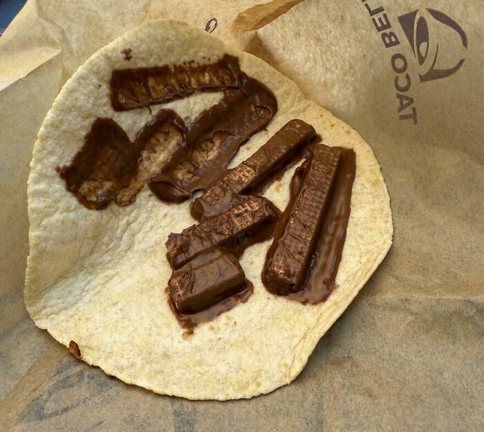 Taco shell filled with melted chocolate pieces on a paper wrapper, an unusual and cursed food combination.