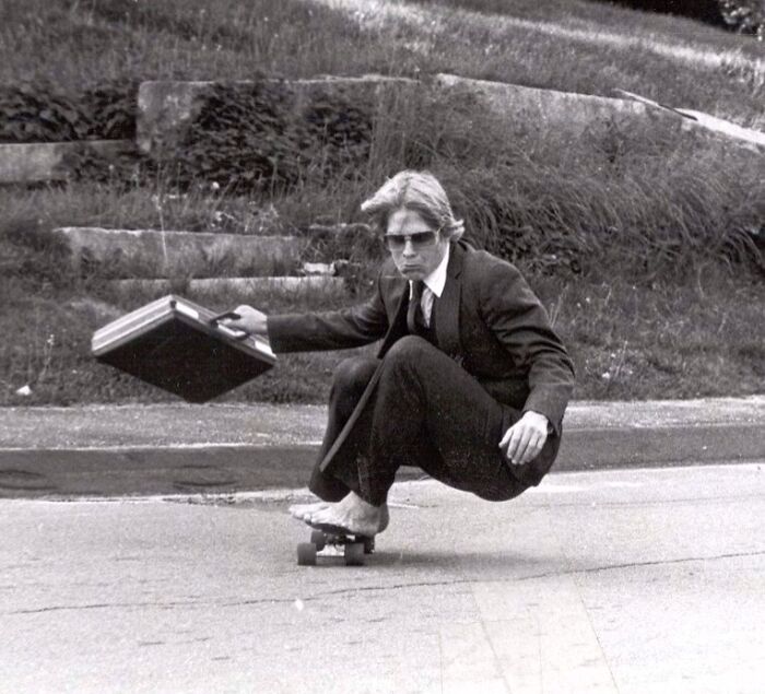 Man in a suit skateboarding with a briefcase on a street, showcasing the wild and weird side of the 1980s.