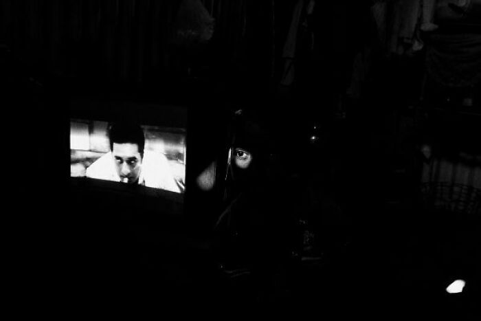 Black-and-white street photo from India showing a dimly lit face beside a glowing television screen.
