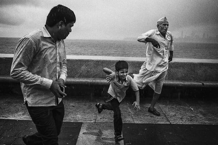 Black-and-white photo of Indian man, elderly person, and child playing along a waterfront street in India.