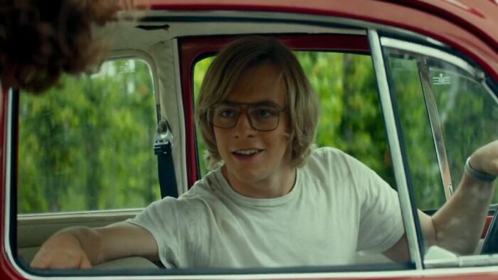 A young man with glasses and blonde hair sits in the driver’s seat of a red car in a movie based on true stories.