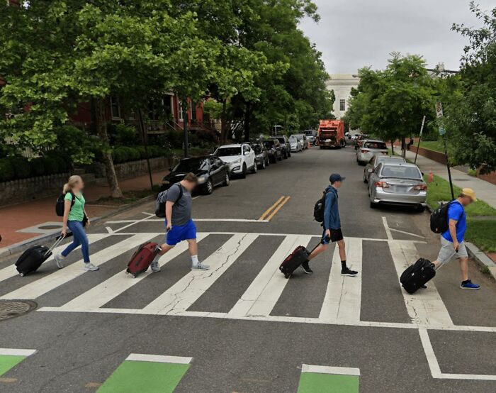 Four people crossing a street with rolling suitcases, captured from an unexpected angle on Google Maps view.