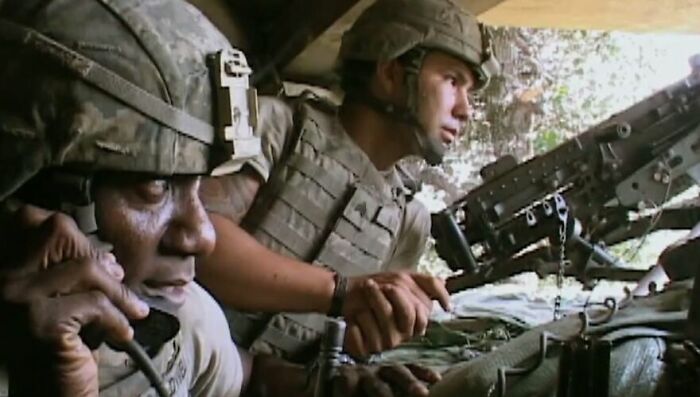 Two soldiers in combat gear operate communications and a mounted machine gun in a war zone, from movies based on true stories.