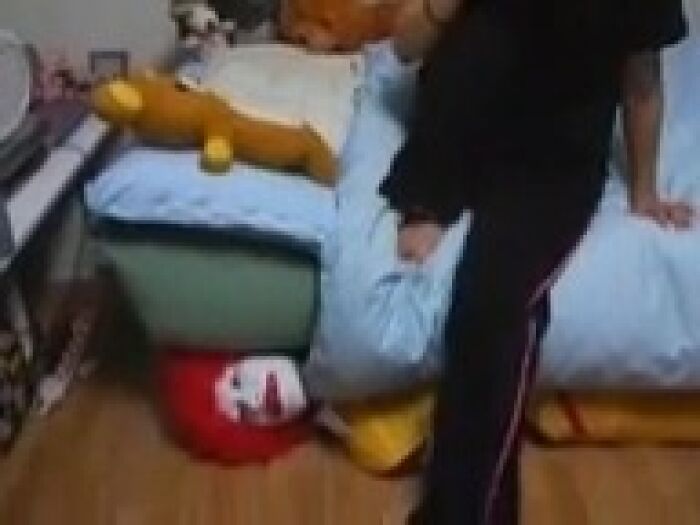 Person standing near a bed with a creepy clown mask partially hidden under the mattress in a dimly lit room.
