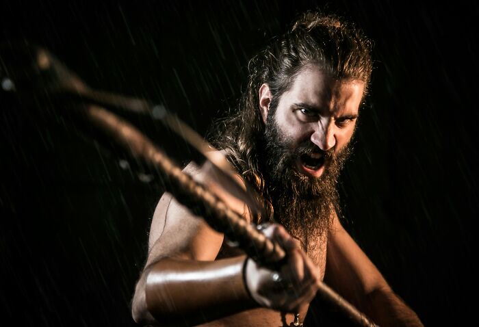 Angry Viking warrior with long hair and beard holding a spear, ready to strike in a dark rainy setting.