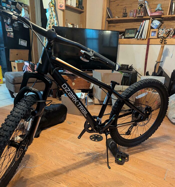 Black Ozark Trail mountain bike missing a front wheel, shown indoors on wooden floor with cluttered living room background.