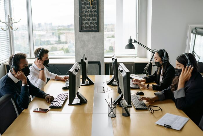 Four office workers wearing headsets and masks, sitting at computers, illustrating workplace rules in a modern office setting.