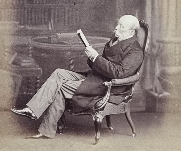 Elderly man in vintage attire sitting in an ornate chair reading a book about famous mysterious imaginary worlds.