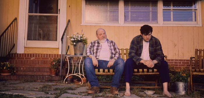 Two men sitting on a wooden bench outside a house, depicting a scene with bad movie details and confusing moments.
