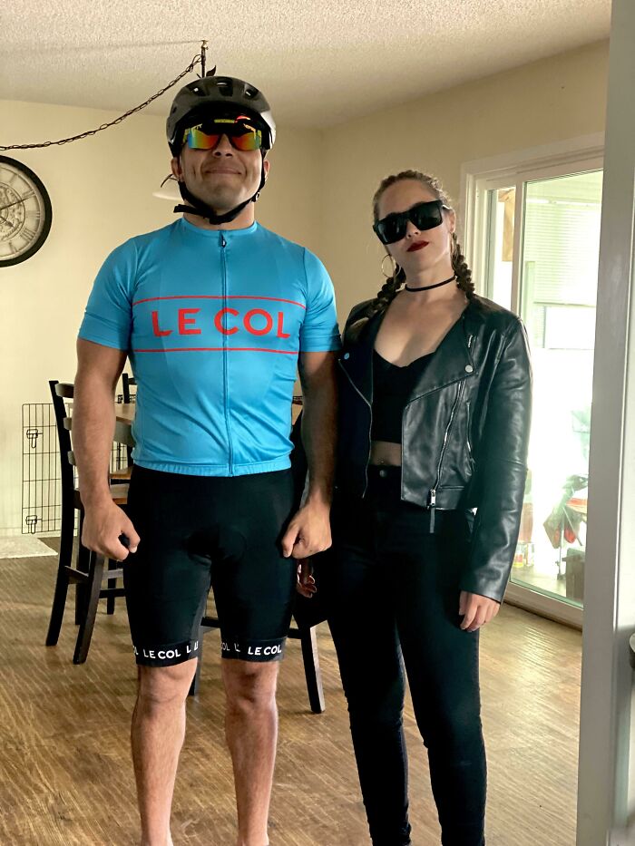 Man in cycling outfit and woman in black leather jacket and sunglasses posing indoors for Halloween fails photo.