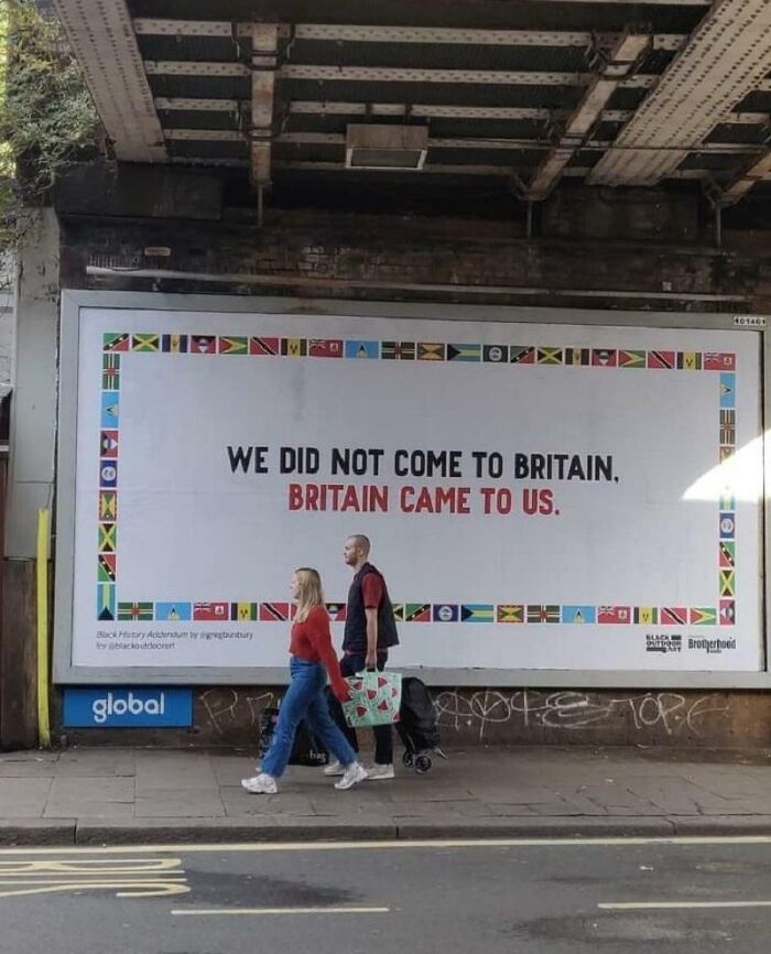 Powerful street art billboard with message about Britain and its history, framed by vibrant international flags in urban setting.