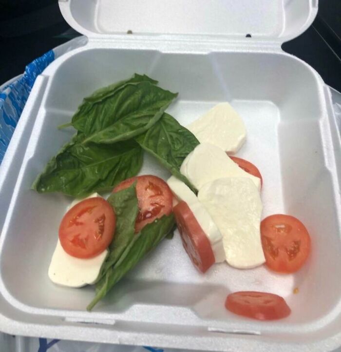 Poorly arranged cursed food with sliced tomatoes, basil leaves, and mozzarella cheese in a white takeout container.