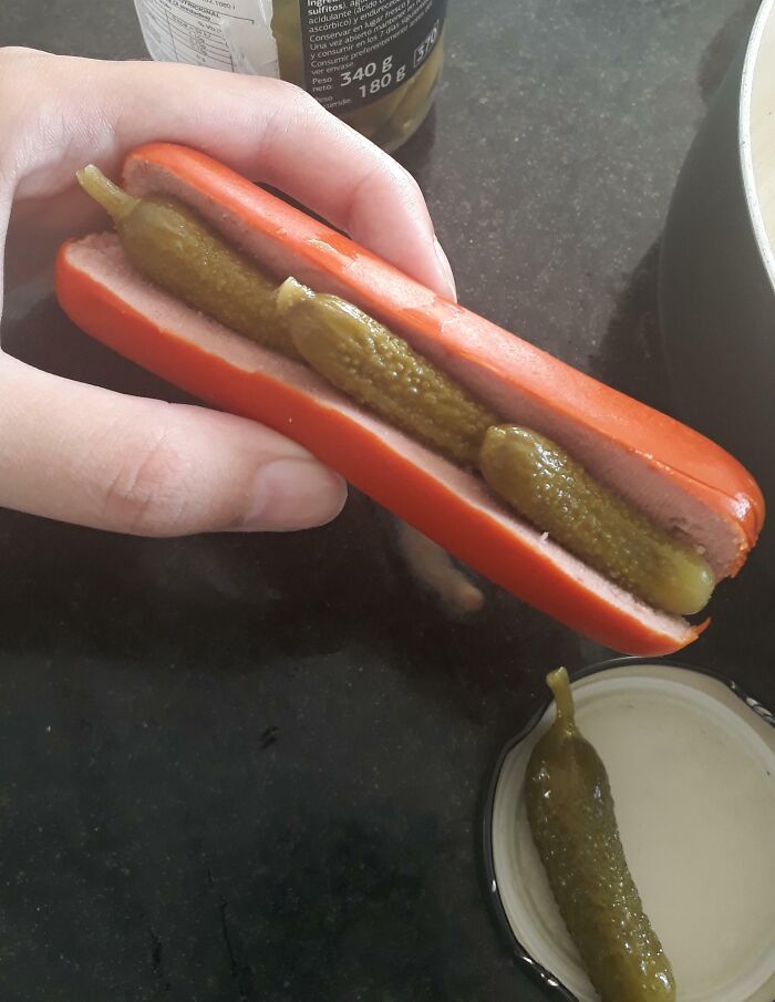 Hand holding a hot dog bun with a sausage filled with whole pickles, a cursed and disgusting food combination.