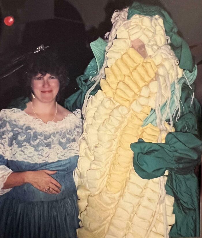 Woman in vintage dress poses next to person in elaborate corn costume, capturing wild and weird 1980s fashion moment.