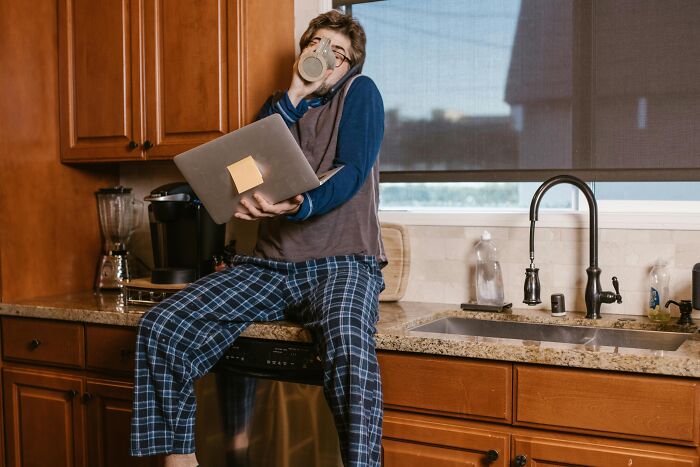 Person in pajamas drinking from a mug while using a laptop in kitchen, illustrating over-the-top workplace rules annoyance.