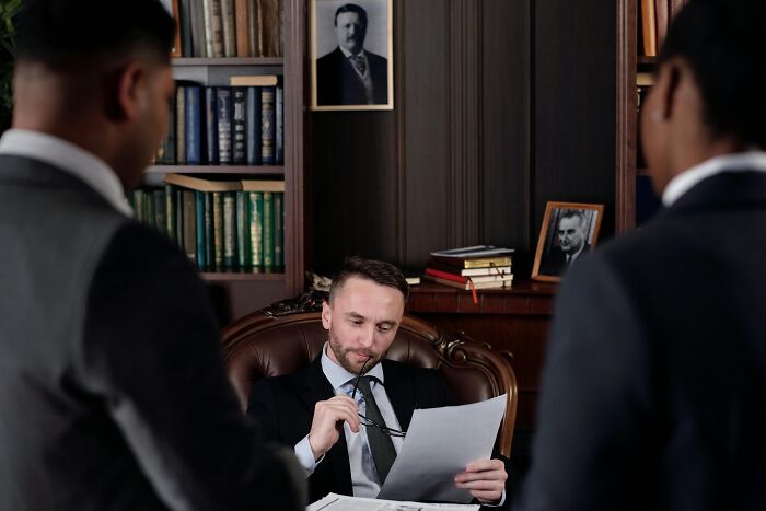 Man in suit reading documents at desk while two colleagues stand nearby in an office highlighting over-the-top workplace rules.