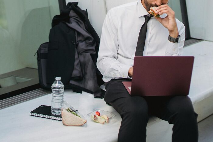Man in business attire eating lunch while working on laptop, illustrating over-the-top workplace rules causing annoyance.