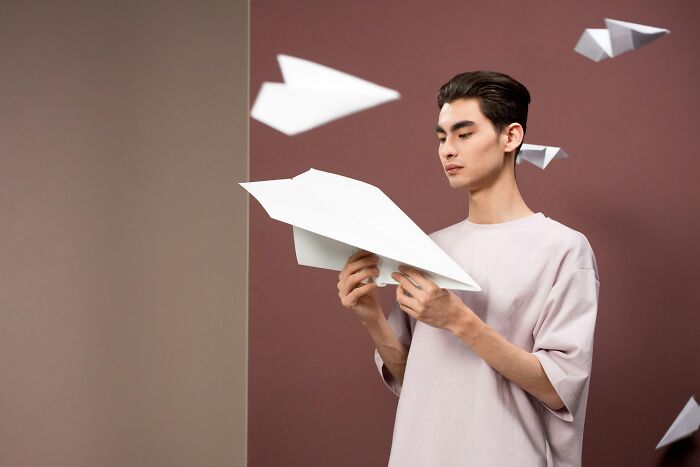 Young man indoors holding a large paper airplane with paper airplanes flying around, illustrating workplace rules annoyance.