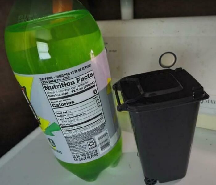 Small black trash bin mistaken for a large soda bottle next to a green soft drink on a kitchen counter.