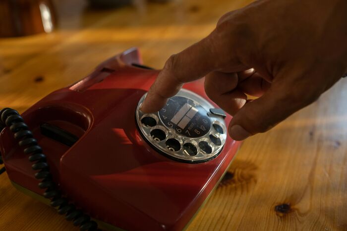 Hand dialing a red rotary phone on a wooden table illustrating over-the-top workplace rules causing frustration.