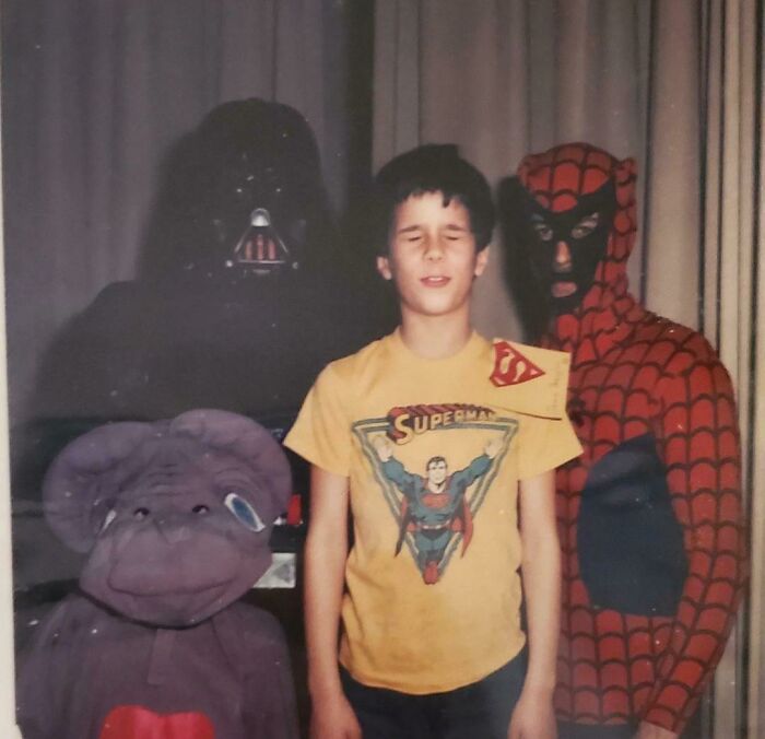 Boy in vintage Superman shirt posing with people in Darth Vader, Spiderman, and alien costumes in a wild 1980s photo.
