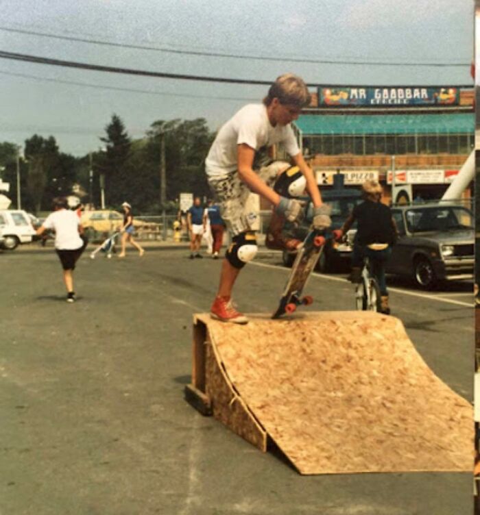 Boy in 1980s gear performing a skateboard trick on a ramp in a busy street scene with bikes and pedestrians.