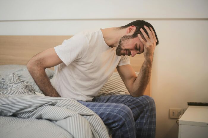 Man sitting on bed in pajamas holding his head, reflecting stress from over-the-top workplace rules causing annoyance.